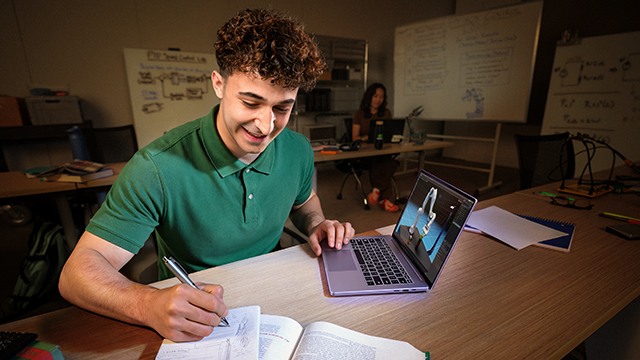 Student przy biurku robi notatki, obok laptop z modelem 3D na ekranie