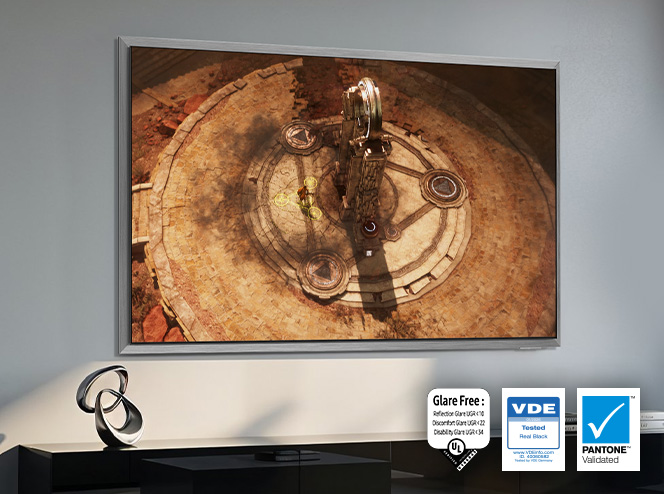 Telewizor Samsung wiszący na jasnej ścianie w mocno oświetlonym pomieszczeniu, na ekranie scena z gry przygodowej ze starożytną kamienną areną. Obok ekranu certyfikaty Glare Free, VDE Tested Real Black i Pantone Validated.